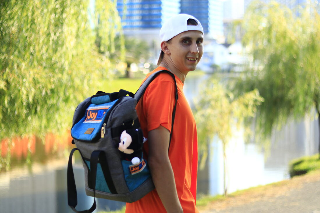 Roman Podvalny in an orange JoyMail courier shirt, carrying a delivery backpack by a river in Batumi.