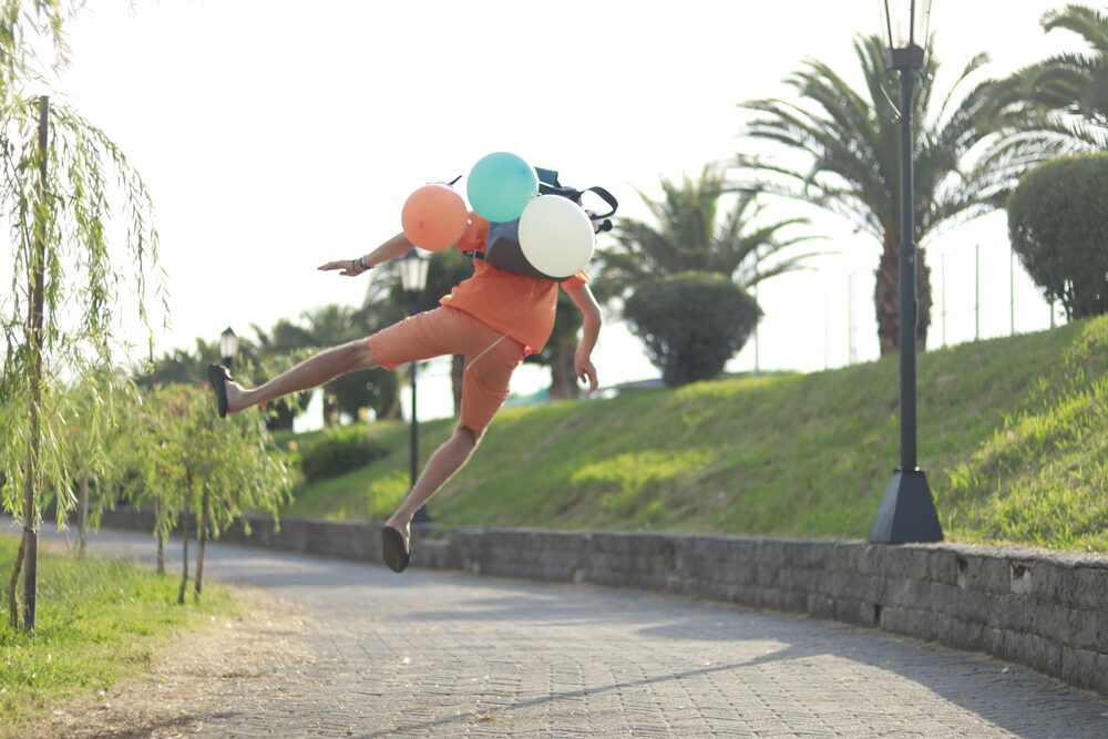 Roman jumping in mid-air on a seaside path, holding a bunch of balloons.