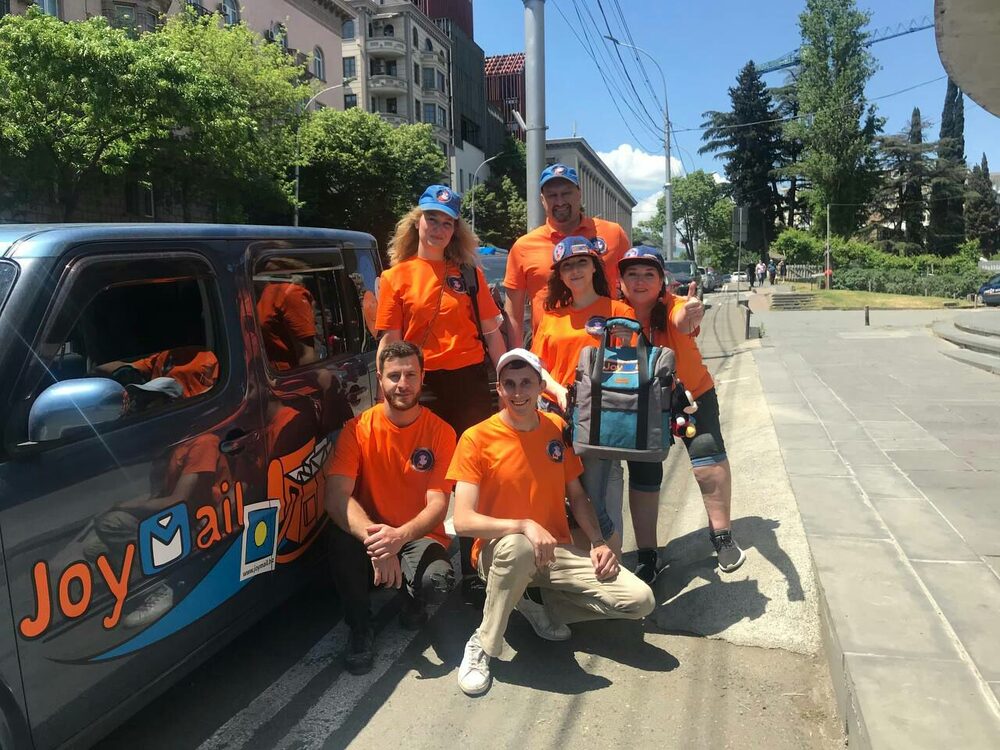 The JoyMail team in orange uniforms posing around a branded delivery van on a street in Georgia.